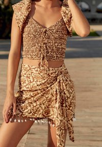 Donna che indossa un crop top a motivo beige e marrone e una gonna a rap sullo stesso tema con frange a conchiglia, in piedi all'aperto su una superficie illuminata dal sole.