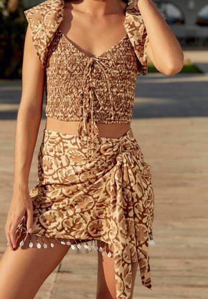 Donna che indossa un crop top a motivo beige e marrone e una gonna a rap sullo stesso tema con frange a conchiglia, in piedi all'aperto su una superficie illuminata dal sole.