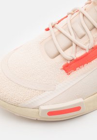Light peach athletic shoe featuring a textured mesh upper, soft fabric accents, coral detailing, and a reinforced sole with a contrasting red stripe.