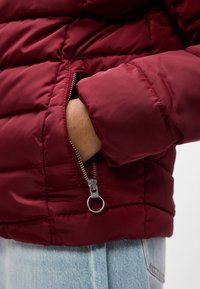 Χέρι εν μέρει μέσα σε φερμουάρ τσέπης κόκκινης καπιτονέ μπουφάν που φοριέται πάνω από ανοιχτόχρωμο μπλε τζιν.