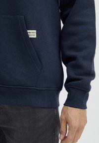 Navyblauwe hoodie van zacht materiaal, met een voorzak, ribbelmanchetten en een label met "PROJECT" en "2020" erop genaaid.