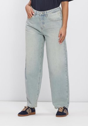 Persoon die losse lichtblauwe spijkerbroek, zwart topje en marineblauwe sneakers met bruine veters draagt, staand met één hand in de zak tegen een effen achtergrond.