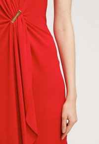 Robe rouge avec un tissu drapé présentant un détail en nœud tordu à la taille, accentué par une pièce en métal. Texture lisse.