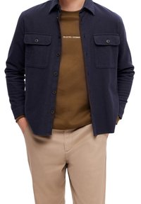 Camisa azul marino abotonada con dos bolsillos en el pecho, hecha de tela texturizada. Se lleva sobre una sudadera marrón con cuello redondo y pantalones beige.