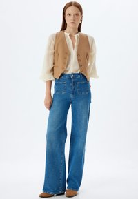 Chaleco beige claro con botones, superpuesto a una blusa blanca, combinado con unos vaqueros de tiro alto y pernera ancha en azul, y zapatos color tostado.