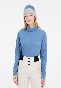 Hellblauer gerippter Rollkragenpullover kombiniert mit hoch geschnittenen cremefarbenen Hosen mit einem schwarzen Bund und drei Knöpfen. Graue Strickmütze auf dem Kopf.