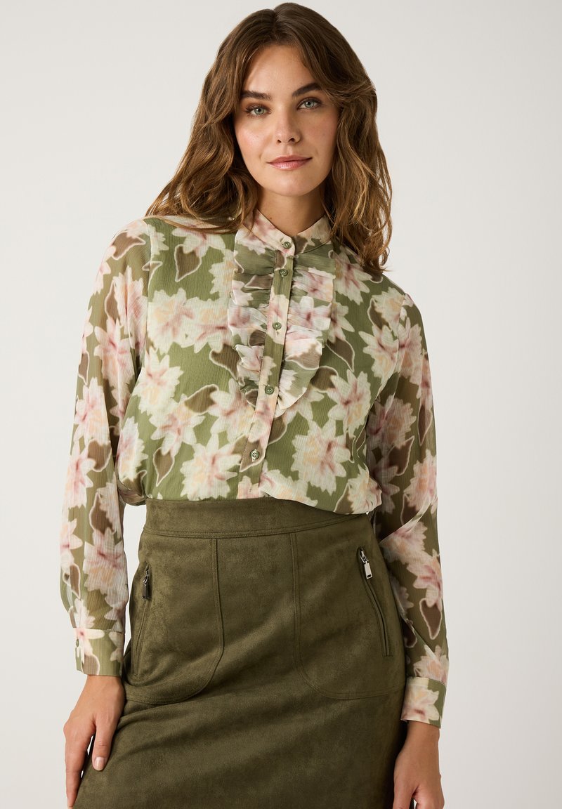Chemisier floral à boutons en tissu vert transparent avec des motifs de fleurs roses, doté d'un plastron volanté et de manches longues, associé à une jupe verte.