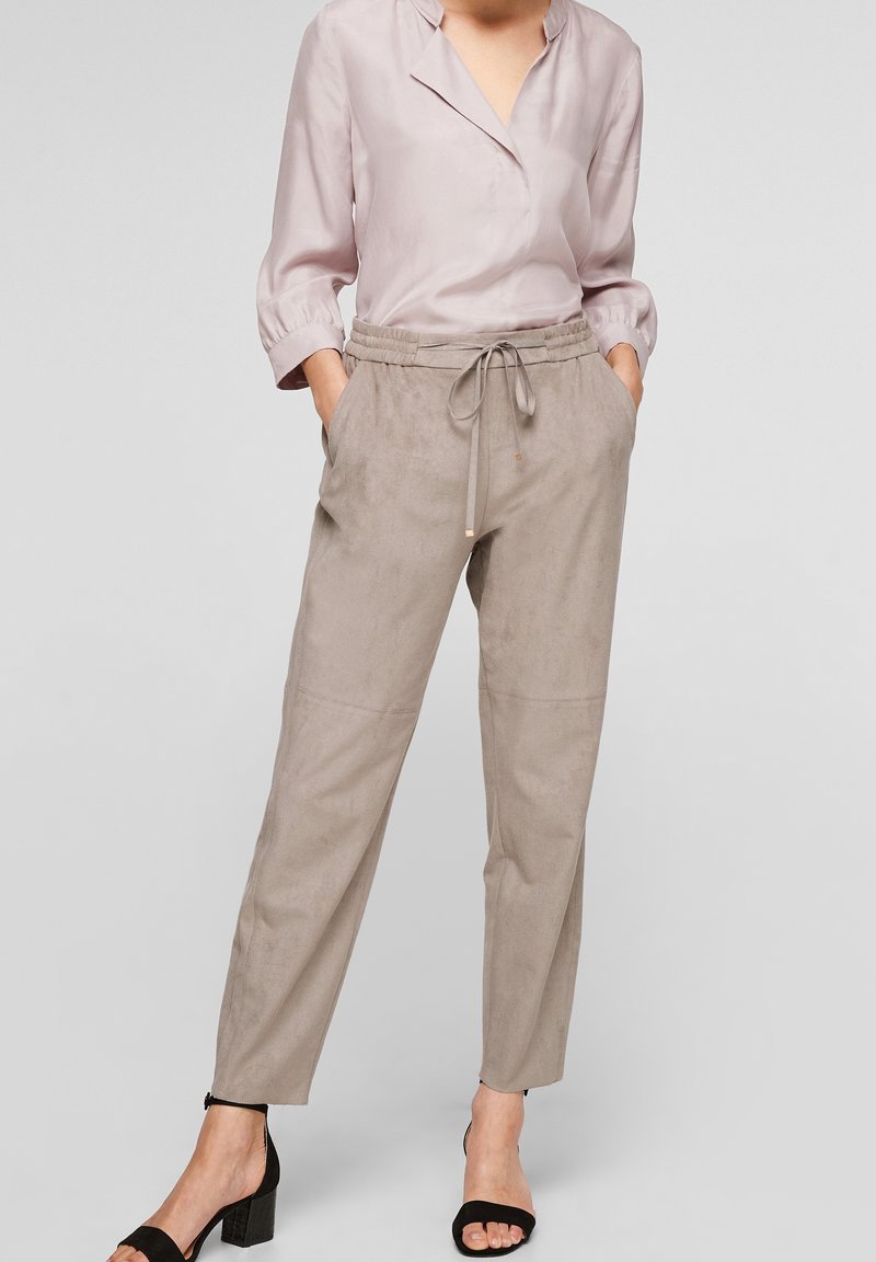 Femme portant un chemisier mauve clair à manches trois-quarts, un pantalon beige à cordon et des sandales noires à talon bloc ouvert, debout les mains dans les poches.