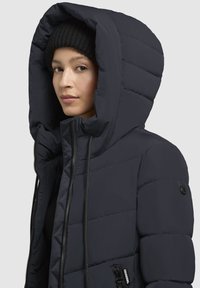 Veste matelassée bleu marine foncé avec une grande capuche, devant zippé et cordons de serrage ; elle présente un tissu texturé et une poche zippée sur le côté.