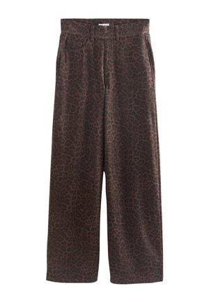 Kvinde med blondt hår iført sort lammeskindjakke, grøn "New York"-t-shirt, brune leopardmønstrede vide bukser og beige sko, stående mod en beige baggrund.
