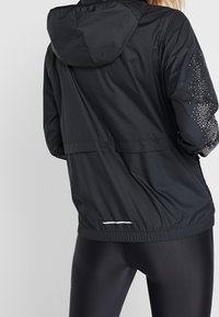 Mujer vestida con una chaqueta con capucha negra y puntos reflectantes en las mangas, y leggings negros ajustados, vista desde atrás.