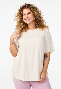 Jasnobeżowy top z krótkim rękawem i okrągłym dekoltem o miękkiej fakturze. Połączony z jasnofioletowymi spodniami. Na ramieniu widoczny charakterystyczny tatuaż.