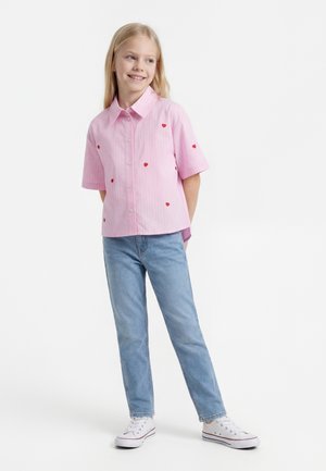 Fille souriante debout, les mains derrière le dos, portant une chemise rose à boutons avec des cœurs rouges, un jean bleu clair et des baskets blanches.