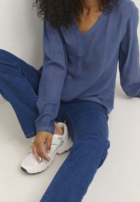 Blaue, langärmelige Bluse mit V-Ausschnitt und strukturierter Oberfläche, kombiniert mit blauen Jeans und weißen Sportschuhen mit grauen Akzenten.