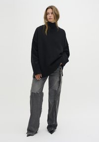 Överdimensionerad svart stickad tröja med hög polokrage, ribbade ärmslut och sidoklyftor, kombinerad med slitna grå denimbyxor med råa kanter.