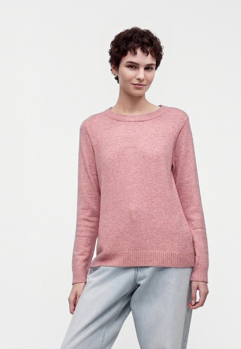 Jeune personne aux cheveux courts et bouclés, portant un pull rose à manches longues et un jean bleu clair, debout devant un fond blanc uni.