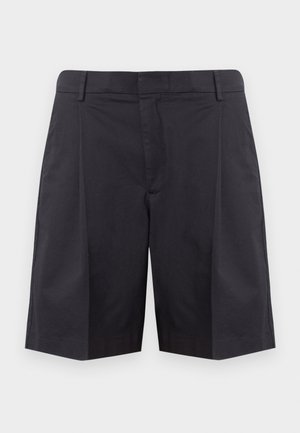 Jeune homme debout les mains dans les poches, portant un haut en maille rayé à manches courtes, un short noir, des chaussettes blanches et des mocassins beiges.