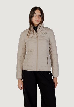 Femme portant une veste matelassée beige avec une fermeture éclair frontale et un col haut, assortie à un pantalon noir, debout devant un fond clair.