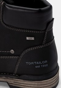 Botín negro de cuero hasta el tobillo con costuras grises, detalles metálicos y una suela en capas. Presenta la etiqueta de la marca "TOM TAILOR" y una etiqueta "TEX" que indica resistencia al agua.