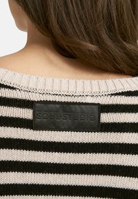 Oberer Rücken einer Frau mit langen braunen Haaren, die einen beigen und schwarz gestreiften Strickpullover mit einem schwarzen Patch trägt, auf dem "SOUL STUDIO" steht.