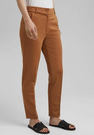 Person trägt schlanke, hoch taillierte braune Hose mit Knopf und Reißverschluss, schwarze Slide-Sandalen und ein weißes Oberteil vor einem schlichten Hintergrund.