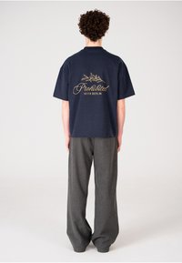 Camiseta de manga corta azul marino con texto dorado bordado y diseño de hojas en la parte posterior, combinada con pantalones grises holgados.