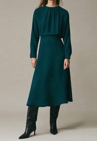 Donna che indossa un vestito midi a maniche lunghe verde scuro con scollatura arricciata e stivali neri con tacco al ginocchio, in piedi davanti a uno sfondo neutro.