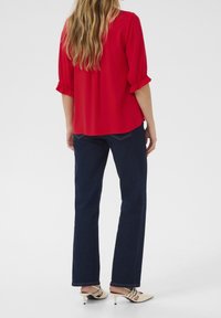 Blusa rossa con maniche a volant abbinata a jeans dritti blu scuro e scarpe con tacco beige con dettagli a cinturino, vista da dietro.