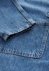 Keskmise sinise varjundiga denimkangas, millel on volditud serv ja tasku, millel on õmblustooted. Tekstuuriga pind, millel on kerge kulunud välimus.
