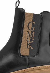 Bottines Chelsea en cuir noir avec un panneau élastique rayé tan et noir portant le logo "GMK". Semelle épaisse et texturée pour une meilleure adhérence.