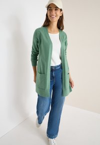 Zelený cardigan s dlouhými rukávy, otevřeným předním dílem a dvěma předními kapsami. V kombinaci s modrými širokými džíny a bílými teniskami.