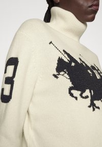 Pull à col roulé crème avec logo joueur de polo noir sur la poitrine et numéro 3 sur la manche gauche, porté par une personne aux cheveux tressés.