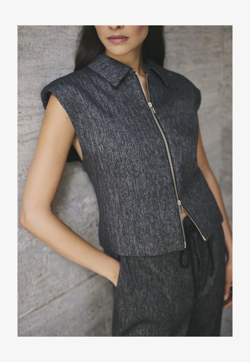 Top din denim gri fără mâneci, cu guler ascuțit și fermoar frontal, având un finisaj texturat. Stilizat cu pantaloni cu șnur, evidențiind o siluetă ajustată.