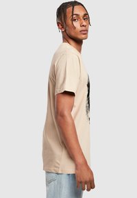 Beige bomulds T-shirt med kortærmer, rund hals, med et sort grafisk design på fronten. Bæres med lyseblå jeans.