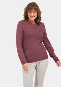 Bordeauxfarbener Strickpullover mit einem strukturierten diagonalen Muster, langen Ärmeln und einem gerippten Rundhalsausschnitt, kombiniert mit hellen Hosen.