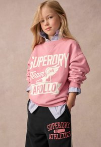 Rosa sweatshirt med vit "SUPERDRY"-logga och vingar grafik, ihop med svarta sweatshirtbyxor med röd text "SUPERDRY ATHLETICS".