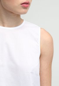 Donna che indossa una blusa bianca senza maniche, labbra leggermente dischiuse, con un tono di pelle neutro su uno sfondo chiaro e uniforme.