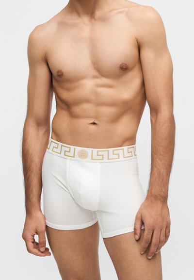 Homme torse nu portant un boxer blanc avec une ceinture dorée à motifs et un emblème, debout devant un fond clair uni.