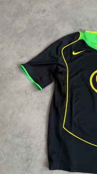 Svart sportstrøye med grønn krage og ermebesetning, gul piping og en gul Nike-swoosh på en grå overflate.