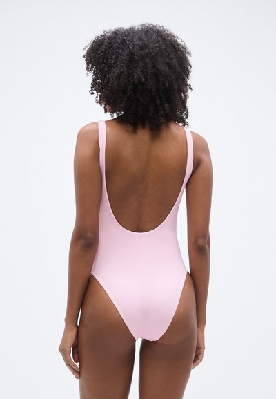 Ροζ ολόσωμο μαγιό με βαθύ ντεκολτέ στην πλάτη, φαρδιές τιράντες και ομαλή, ματ υφή. Σχεδιασμένο με μινιμαλιστικές γραμμές και καμπύλες.