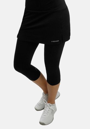 Parte inferiore del corpo con leggings neri fino al ginocchio, una gonna nera e scarpe da ginnastica bianche, in piedi con una gamba incrociata sull'altra.