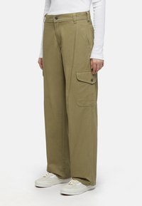 Licht olijfgroene cargo broek met wijde pijpen, zijk anten en een knoopsluiting, gemaakt van stevige stof. Gepaard met witte sneakers.