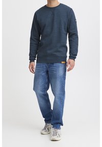 Sudadera azul marino de mangas largas, cuello redondo y tejido texturizado; combinada con vaqueros de mezclilla azul claro y zapatillas beige.