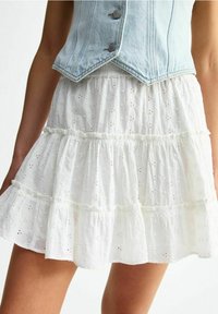 Jupe blanche à volants en tissu léger avec des motifs brodés et des bords festonnés. Portée avec un gilet en jean clair.