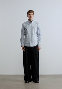 Ανοιχτό μπλε πουκάμισο με κουμπιά, μακριά μανίκια, τσέπη στο στήθος με λογότυπο, σε συνδυασμό με άνετο μαύρο παντελόνι, φορεμένο με μαύρα παπούτσια.