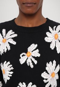 Stickad svart tröja med vita prästkragar med orangea mittpunkter. Texturerat tyg och ribbad halsringning. Blommönster över framsidan.