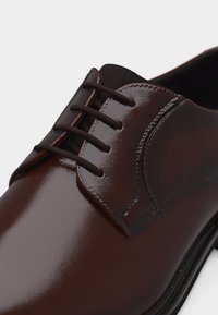 Καφέ γυαλιστερό δερμάτινο παπούτσι επίσημου τύπου με κλειστό κορδόνι και λεπτά στρογγυλά σκούρα καφέ κορδόνια, σε απλό ανοιχτό φόντο.