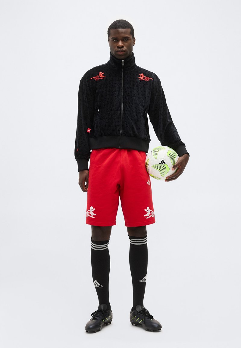 Man staat met een witte en groene Adidas voetbal, draagt een zwart jack met ritssluiting, rode shorts met witte logo's, zwarte Adidas sokken en zwarte voetbalschoenen.