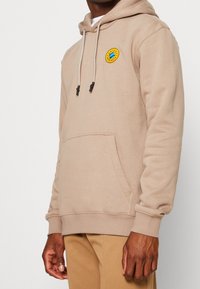 Beige luvtröja med ficka fram, dragsko och en rund, färgglad logotypapplikation på bröstet. Mjukt tygmaterial.