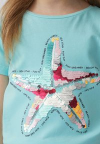 Lichtblauwe katoenen t-shirt met een groot zeesterontwerp van multicolore sequins, met de tekst "SUN DREAMER," "BEACH VIBES," en "FUN DAY."